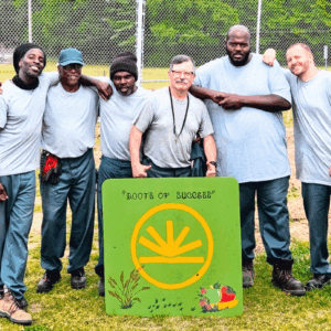 RoS Garden - square Program participants standing together outdoors behind a hand-painted Roots of Success sign.